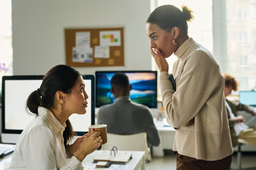 Wall Mural - Two intercultural female office managers gossiping at coffee break while one of them covering mouth by hand and looking at colleague