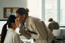 Young Smiling Black Woman With Tablet Bending Over Female Colleague With Ponytail While Whispering Something To Her Ear In Office