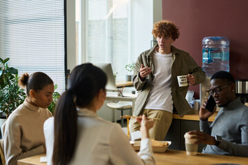 Young businessman with cup of tea or coffee standing by water cooler in front of intercultural colleagues and telling them something