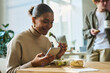 © pressmaster - Happy young African American businesswoman watching video in smartphone and having salad at lunch break in office
