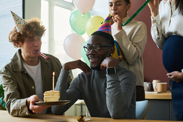 Poster - Happy young man in birthday cap looking at piece of tasty cake with burning candle while sitting by table among colleagues