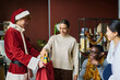 Young smiling African American businesswoman taking giftbox out of red sack held by male colleague wearing Santa Claus costume