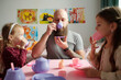 Young bearded father with toy plastic cup and saucer having tea with his two adorable daughters while sitting by small pink table