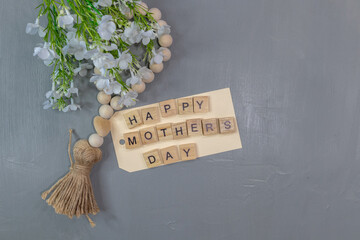 Wall Mural - Happy Mothers Day word tiles on gray background with wood tassel and white flowers flat lay