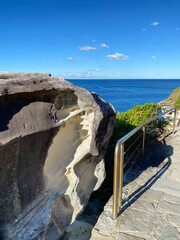 Wall Mural - Big rock, metal fence and bridge over the sea. Boardwalk near the blue ocean. Rock formation under a blue sky. Rocky cliff. Coastal landscape.