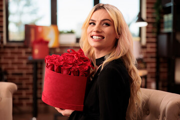 Wall Mural - Cute woman holding valentine day roses in heart shaped box, standing in living room filled with romantic presents. Seductive girlfriend enjoying celebrating love holiday, gifts and flowers bouquet