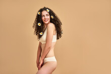 Self Care. Sensual Caucasian Lady Dressed In Only Underwear Looking Aside While Posing Over Beige Background. Young Skinny Woman Wearing Chamomile Bloom In Curly Dark Hair In Studio. Copy Space.