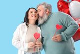 Fototapeta Panele - Beautiful mature couple with heart-shaped balloons and paper hearts kissing on blue background. Valentine's Day celebration
