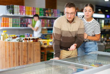 Sticker - Positive interested man shopping together with adult daughter in supermarket, choosing foods in glass refrigerated display case..