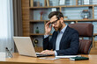 © Liubomir - Frustrated administrator experiencing headache while sitting by desk with portable computer in modern interior. Exhausted man feeling pain in temple and getting distracted from working process.