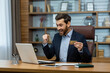 © Liubomir - Excited brunette man looking at portable computer screen and making winner gesture while holding bank card in office. Joyful collector purchasing valuable item at online auction with cashless payment.