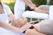 © Elliott/peopleimages.com - Women, hands and massage with hot stone in spa, outdoor and wellness on bed for body treatment. People, heat and masseuse for release of muscle tension, nature and health detox in comfort at resort