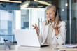 © Liubomir - Senior smiling gray-haired woman in a suit and headset works in a call center, sits in the office at a laptop, consults and conducts an online video meeting, smiles and gestures with her hands.