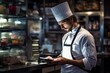 © Iftikhar alam - A man wearing a chefs hat focuses on a tablet as he seeks culinary inspiration in a busy professional kitchen, Restaurant chef orders groceries to kitchen using tablet, AI Generated