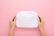 Young adult woman hands holding and showing new white cosmetic bag on light pink table background. Pastel color. Closeup. Point of view shot. Top down view.