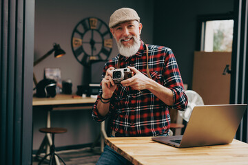 Wall Mural - Portrait d'un homme photographe hipster situé dans un studio atelier et qui tient un appareil photo vintage en souriant devant un ordinateur