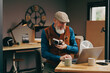 Homme photographe assis souriant quinquagénaire senior hipster élégant et stylé qui travaille sur un ordinateur dans un atelier créatif vintage et qui tient un appareil photo en buvant un café