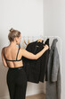 Elegant young woman standing with her back in front of the modern rack with capsule collection of clothing and holding a black jacket. Modern consumer concept and lifestyle.