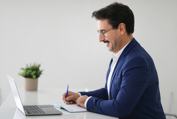 Wall Mural - Cheerful caucasian adult businessman in suit at table with laptop, make notes