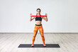 Portrait of strong sporty woman wearing black tank top and orange leggings, doing workout with latex resistance band, training her arms. Full length indoor shot against white wall.