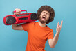 © khosrork - Portrait of excited man with Afro hairstyle wearing orange T-shirt listening to music holding red tape recorder on shoulder, showing rock and roll sign. Indoor studio shot isolated on blue background.