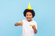 © khosrork - Portrait of birthday little boy with curly hair wearing yellow party cone holding tasty delicious donut with candle showing thumb up like gesture. Indoor studio shot isolated on blue background.