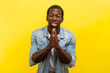 Portrait of pleading man standing with palms together praying having problems asking to forgive apologies, wearing denim casual shirt. Indoor studio shot isolated on yellow background.
