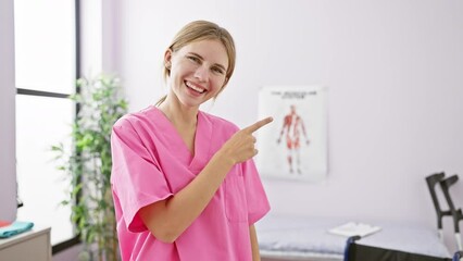 Wall Mural - Cheerful, beautiful blonde girl, working hard at rehab clinic, smiling confidently and optimistically as she points with her hand and finger towards the side.