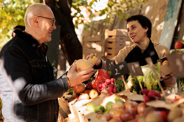 Wall Mural - Friendly young farmer selling organic vegetables from farming stand at farm market, talking to customer. Local female vendor showing eggplant to senior man buying fresh and healthy food.