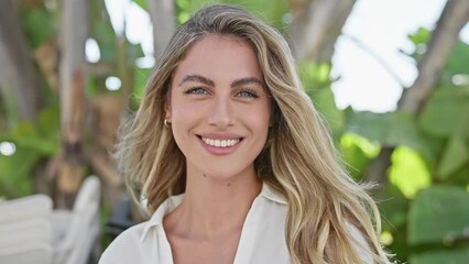 Wall Mural - Cheerful young blonde woman exuding confidence, joyfully smiling while standing in a sunny park, reveling in the green beauty of nature - a radiant portrait capturing her contagious happiness.