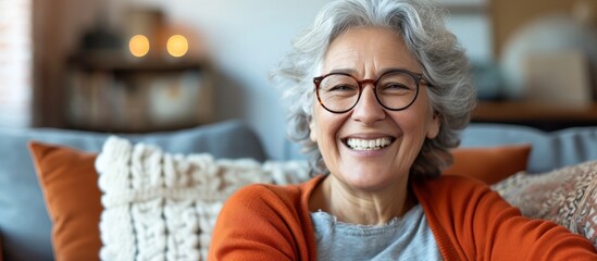 Cheerful older woman in glasses, smiling and laughing on sofa, displaying her healthy teeth while enjoying leisure at home.