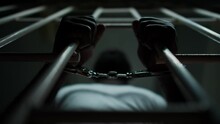 Directly Below View Of Hands Of African American Inmate In Handcuffs Holding Bars Of Prison Cell
