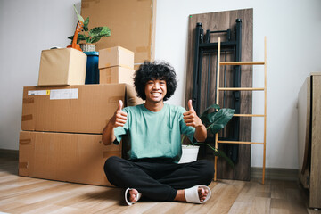 Cheerful young Asian guy sitting cross legged on floor and showing two thumbs up at camera with pile of carton boxes on the background