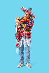 Wall Mural - Young Mexican man in sombrero and with painted skull on blue background. Celebration of Day of the Dead