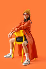 Poster - Stylish young Asian woman sitting in chair on orange background