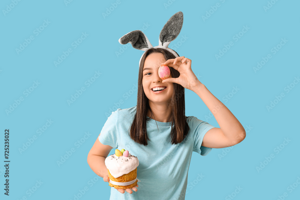 Pretty young woman with bunny ears, Easter egg and cake on blue background