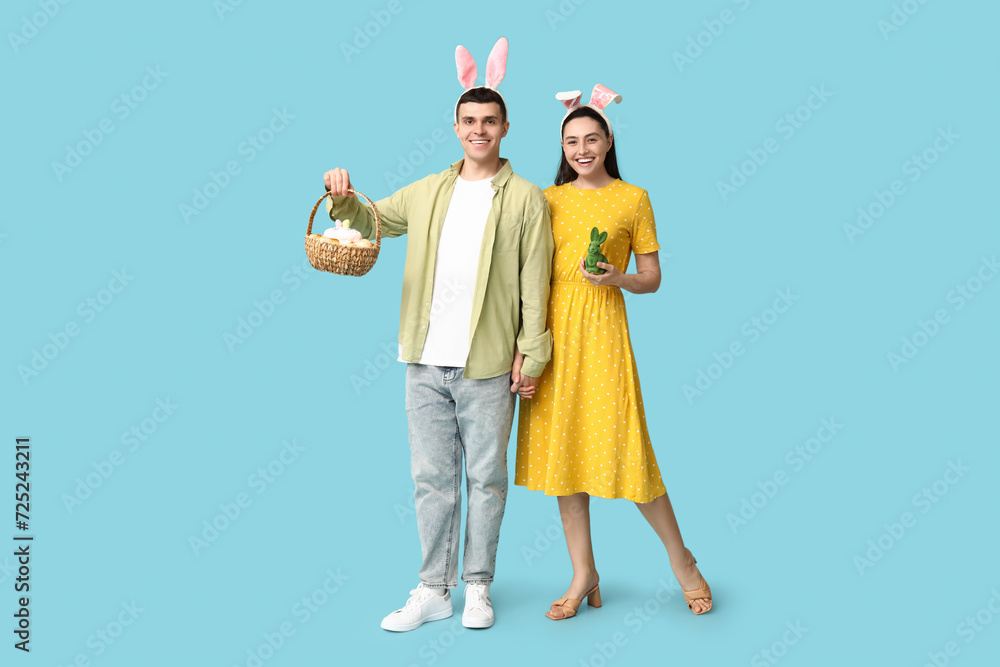 Happy young couple with Easter bunny ears, basket and toy rabbit on blue background