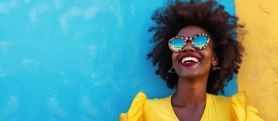 Canvas Print - Joyous black woman with afro curls, dressed stylishly, smiling near a blue wall, radiating confidence and enthusiasm with sunglasses on.