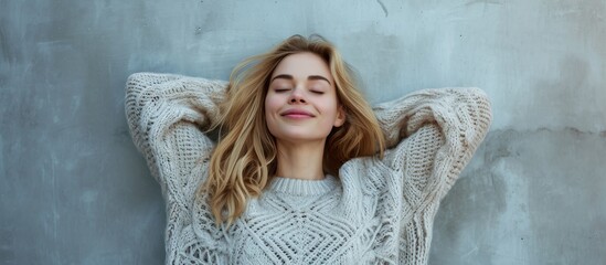 Wall Mural - Blissful blonde woman in a cozy sweater, closing her eyes and stretching her arms on a grey surface.