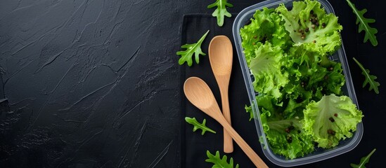 Canvas Print - Top view of a plastic container with a light meal of lettuce, wooden spoons, and green tea on a black background, leaving space for text.