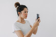 © Suthida - Asian Thai woman using mobile phone with funny doubt face, holding smartphone by silent suspicious, standing isolated over white background wall.