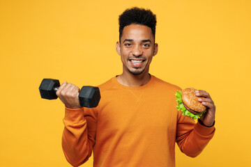 Wall Mural - Young smiling happy man of African American ethnicity wear orange sweatshirt casual clothes eat fast food burger hold dumbbell isolated on plain yellow background studio portrait. Lifestyle concept.