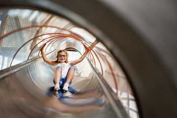 Wall Mural - Little preschool girl riding from childrens slides on playground. Happy smiling child having fun in game center for kids.