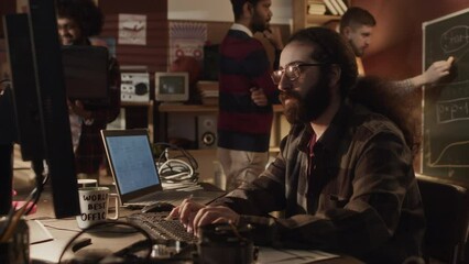 Waist up of long-haired bearded tech lover typing program codes on keyboard while working with other diverse geeks in handmade old-fashioned garage office with warm yellow dimmed light