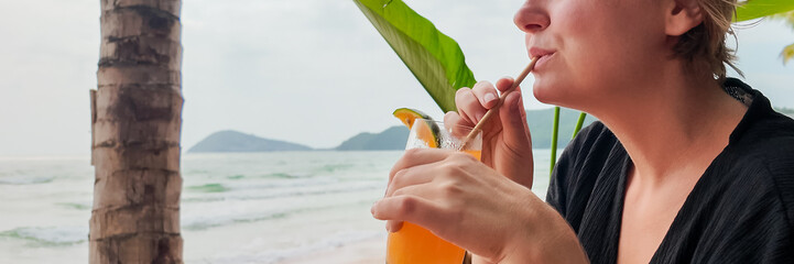 Wall Mural - Woman sipping a tropical drink on a beach with palm trees and ocean backdrop, conveying a vacation or summer relaxation concept