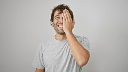 Wall Mural - Confident, cheerful young man with a beard, casually covering one eye with hand and sporting a cheeky smile on his face, against a white isolated background.