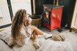 © polinaloves - Young woman sitting by the fireplace in white sweater, drinking tea in cozy log cabin.