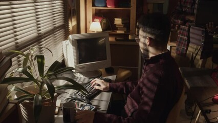 Medium lowkey shot of young Biracial bearded geek sitting at desk in front of window with closed window blinds in dark room and typing program code on old computer