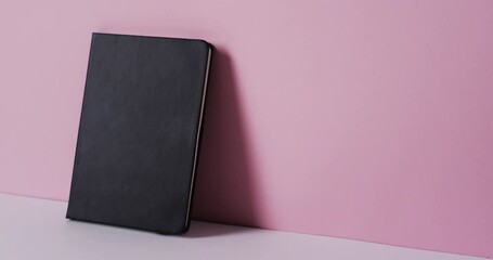 Sticker - Close up of closed black book leaning on wall with copy space on pink background in slow motion