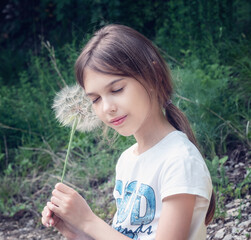 Wall Mural - Closeup portrait of a beautiful little girl on the background of a green field with a big dandelion in her hand